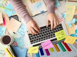 A person working on a laptop at a desk space covered in post-it notes and office supplies is experiencing anxiety.
